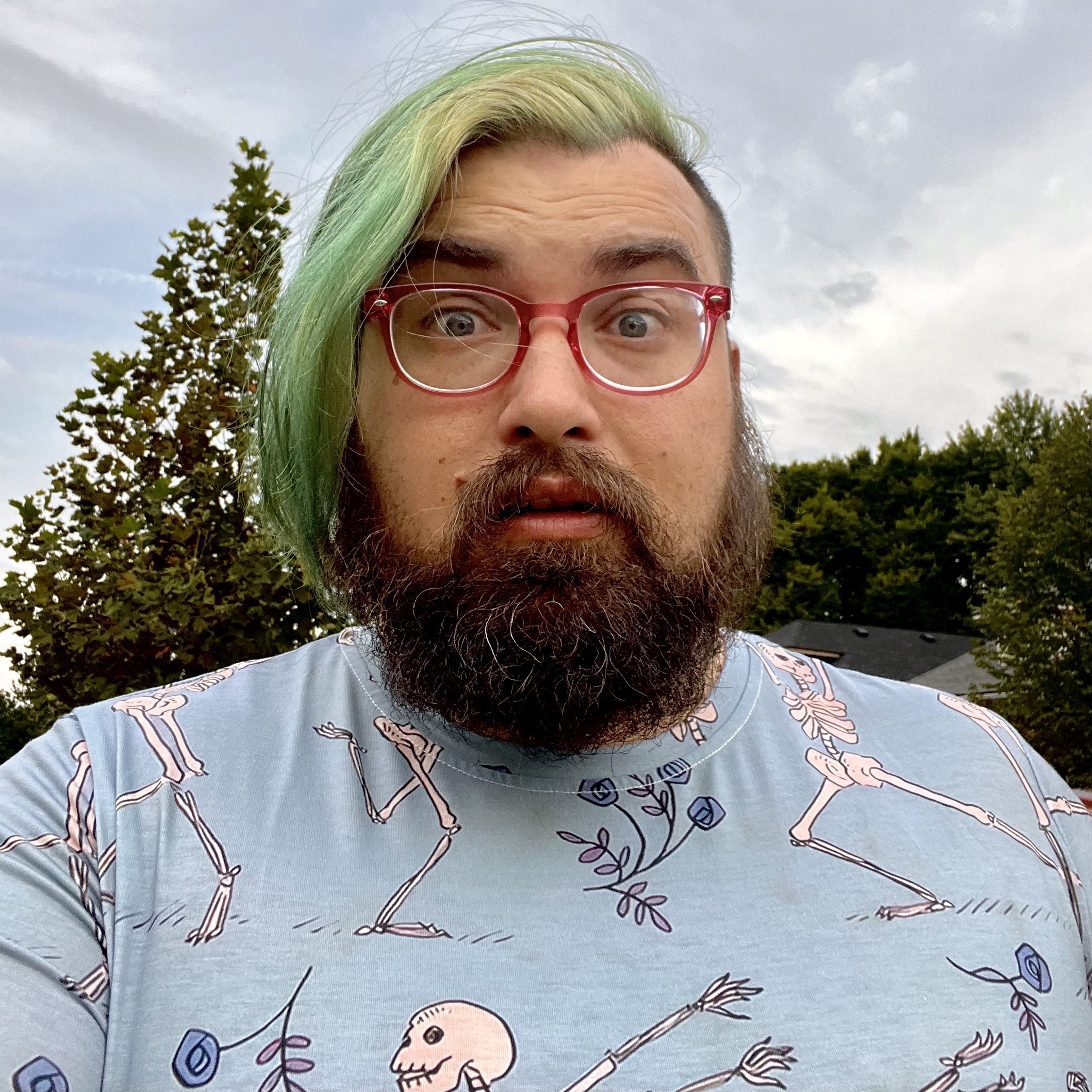 Alex Bezuska (he/him) in a blue shirt with dancing skeletons, standing outdoors in early fall with green to teal dyed hair in an undercut, a medium-sized brown beard with a few greys, and pink-rimmed glasses.